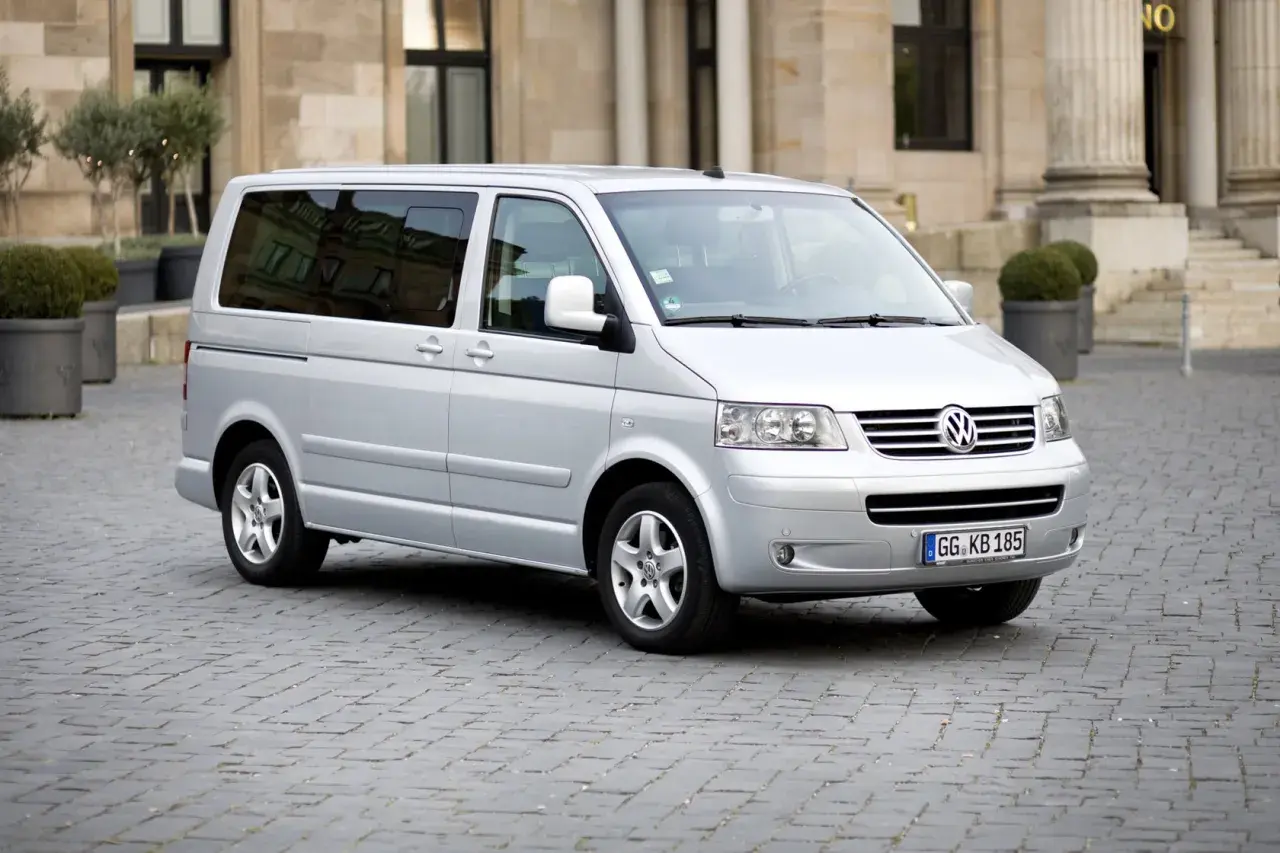 Srebrny Volkswagen T5 na brukowanym placu przed budynkiem z kolumnami.